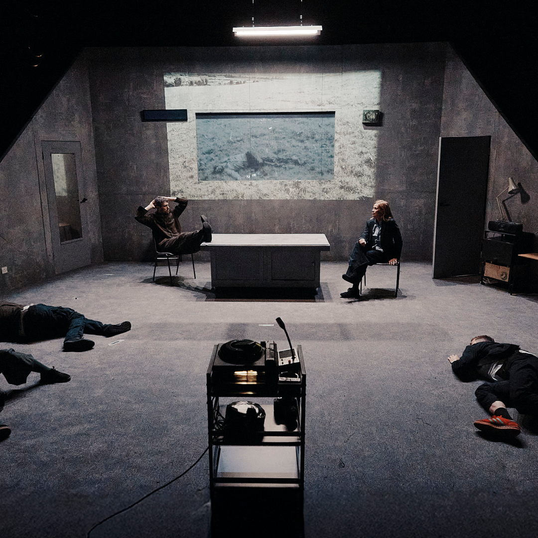 A dimly lit room at Scorched Earth - Brisbane Powerhouse with four people lying on the floor and two at a desk. A projector displays a black-and-white image in the background, adding to the tense, dramatic atmosphere.