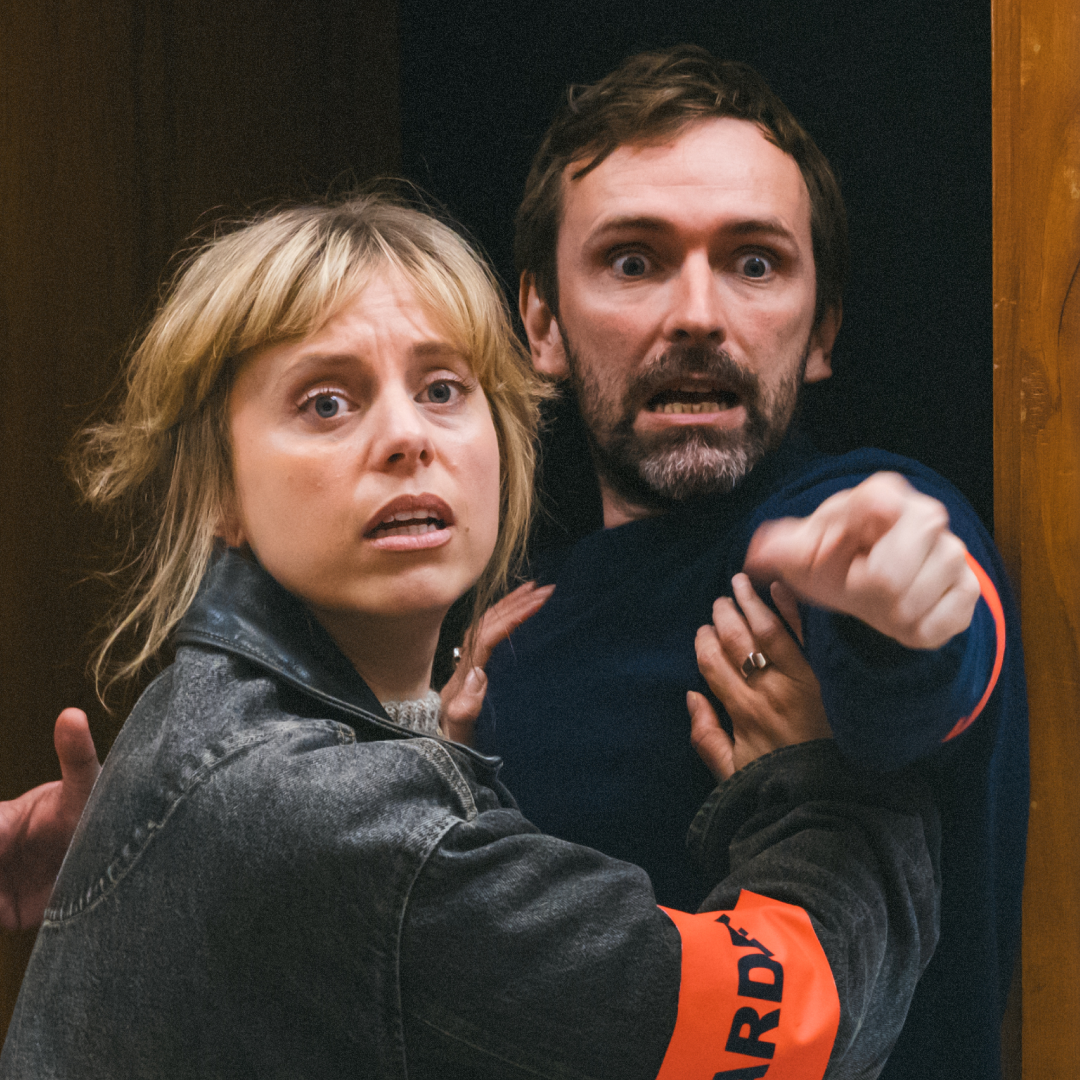 A woman in a denim jacket holds back a concerned man in a navy sweater, both appearing alarmed as they stand in a doorway at Brisbane Powerhouse, their intense expressions suggesting they've just spotted a werewolf.