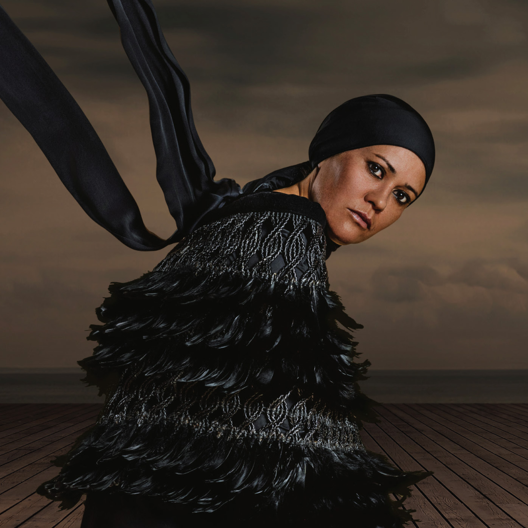 A person wearing a black headscarf and a textured black garment with ruffled layers kneels on a wooden deck against a moody, cloudy sky. A long black scarf flows dramatically in the wind behind them.