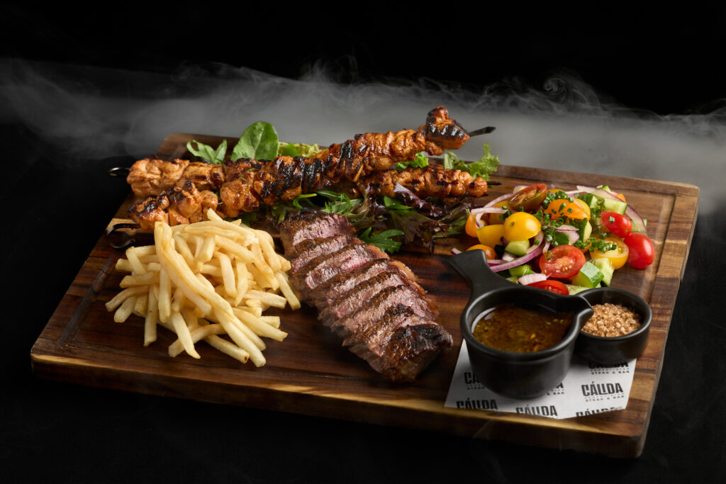 A wooden board with sliced grilled steak, chicken skewers, French fries, fresh salad with cherry tomatoes, a small bowl of sauce, and coarse salt—perfect for sharing at Night Feast 2025 Brisbane Powerhouse, with steam rising in the background.