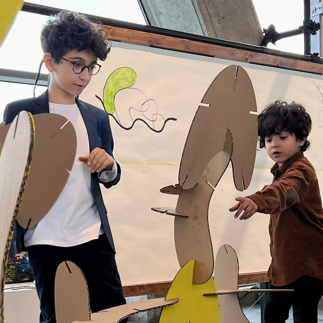Two young boys are building a large structure with interlocking cardboard and yellow pieces. One wears glasses and a blazer, the other a brown shirt. A drawing is visible in the background.