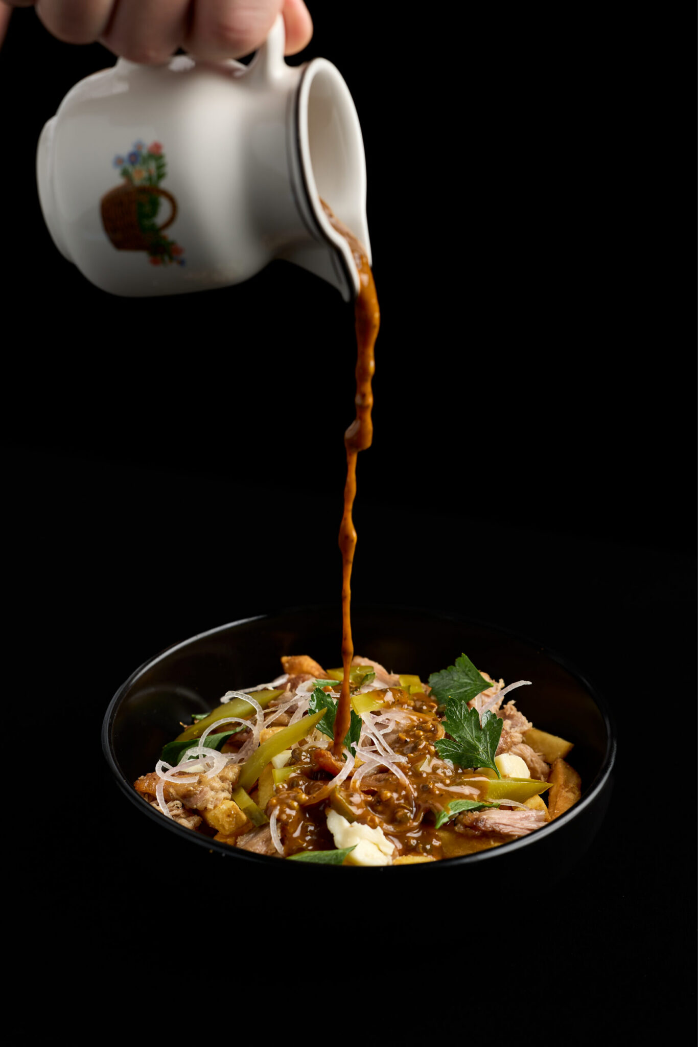 A hand pours brown sauce from a white jug onto a bowl of noodles topped with shredded chicken, vegetables, and herbs, set against a black background—capturing the essence of Night Feast 2026 at Brisbane Powerhouse.