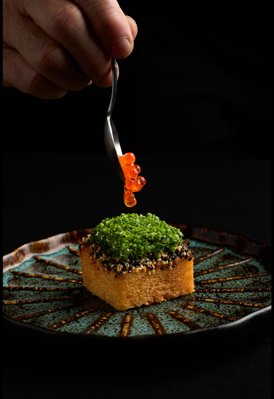 A hand uses a spoon to place orange fish roe onto a square piece of bread topped with green herbs and sesame seeds, served on a decorative ceramic plate—an elegant bite at Night Feast 2026 Brisbane Powerhouse against a dark background.