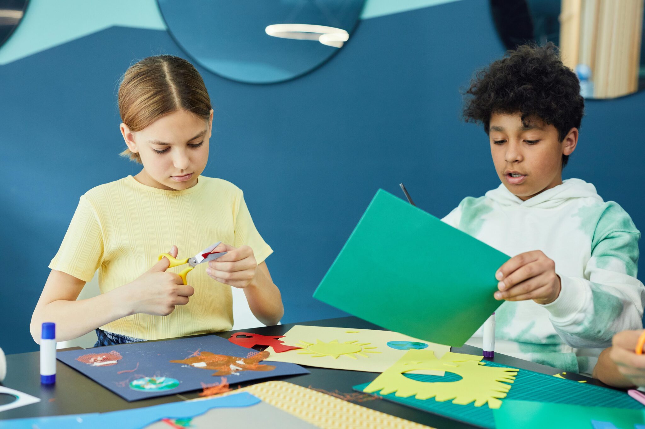Two children sit at a table doing arts and crafts. One girl cuts yellow paper with scissors, while a boy holds a green sheet. Colorful paper, glue sticks, and craft supplies are spread on the table.