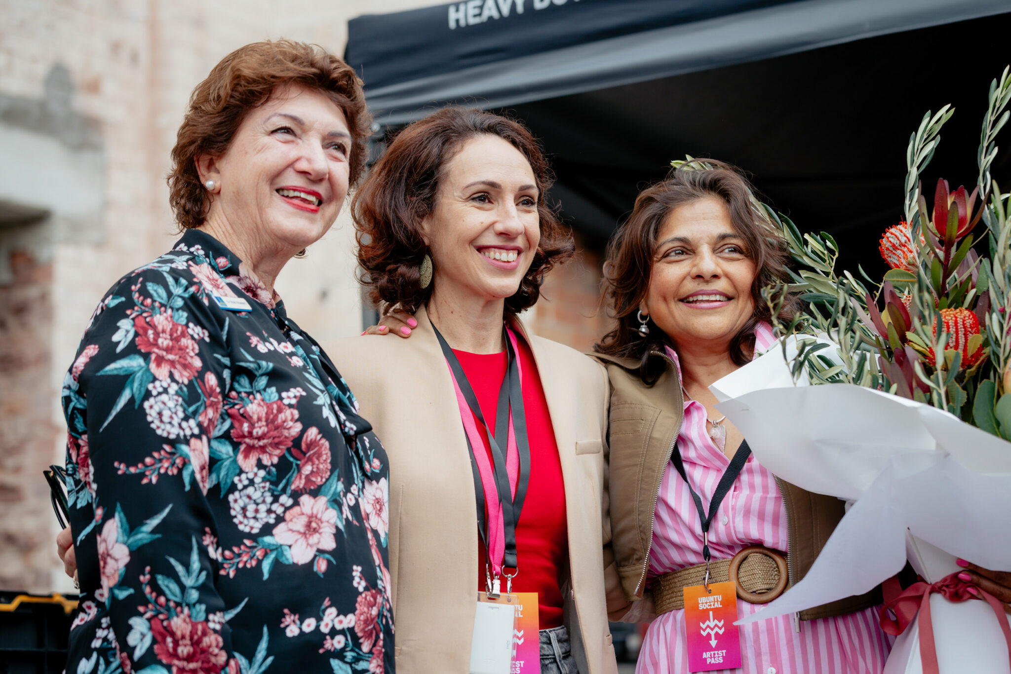 Three women smiling together outdoors, one holding a large bouquet of flowers. They are dressed formally with name badges, standing under a canopy with a brick wall in the background.