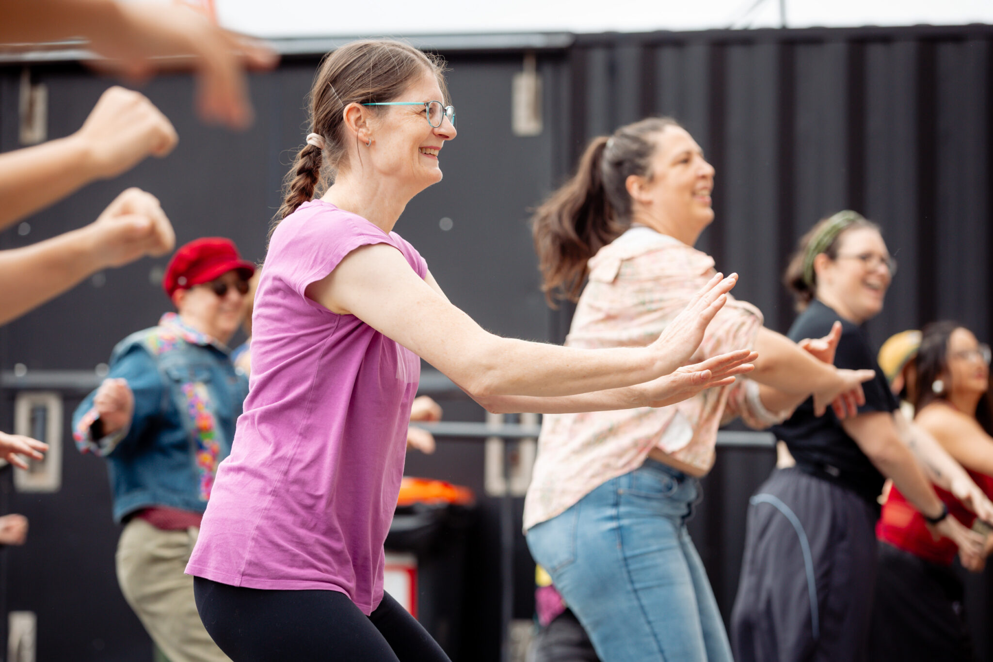 A group of people, smiling and laughing, dance outdoors together. The focus is on a woman in a purple shirt leading the activity, with others mirroring her movements.
