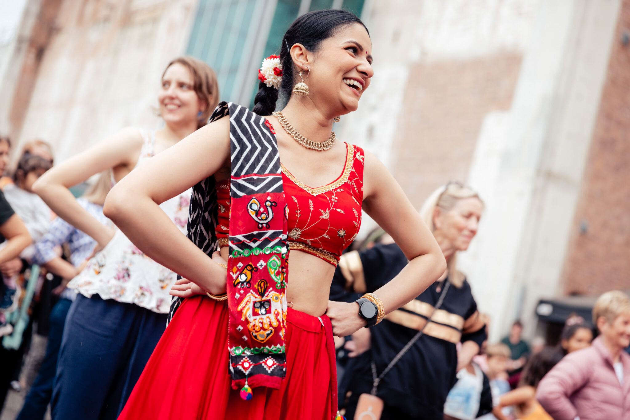 A woman in a red traditional outfit smiles with hands on her hips during an outdoor event. Others around her also pose cheerfully, suggesting a festive and lively atmosphere.