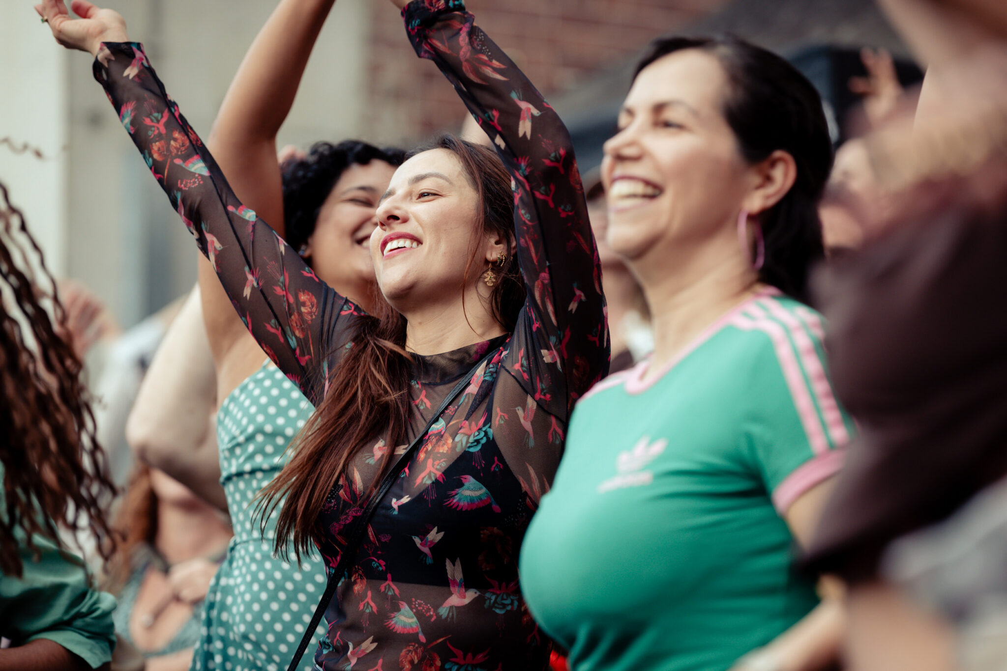 A group of women smiling and cheering outdoors, with one woman in a sheer black top raising her arms joyfully among the crowd.