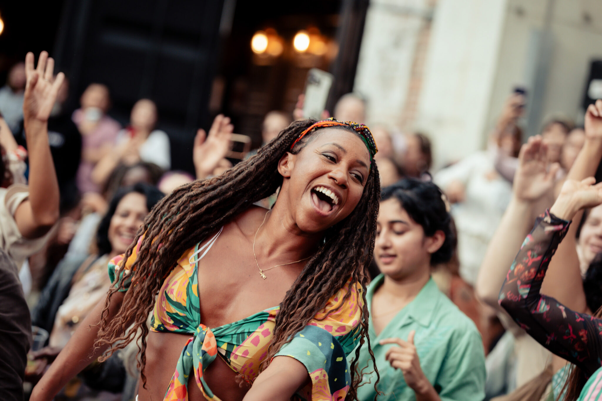 A woman with long braids in a colorful outfit joyfully dances in a lively crowd outdoors, smiling with her arms raised. Others around her are also dancing and celebrating.