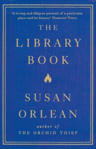 Blue book cover with gold text: The Library Book by Susan Orlean, author of The Orchid Thief. A review quote from Financial Times appears at the top. A small flame icon is beneath the title.