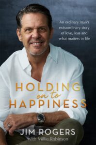 Smiling man in a white shirt sits against a dark background. Text reads: An ordinary mans extraordinary story of love, loss and what matters in life. Holding on to Happiness. Jim Rogers with Millie Robinson.
