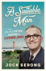 Book cover for A Suitable Man by Jock Serong, featuring a smiling man in glasses and suit, a building labeled Fletcher Jones & Staff, and a large white sphere on a red pole in the background.