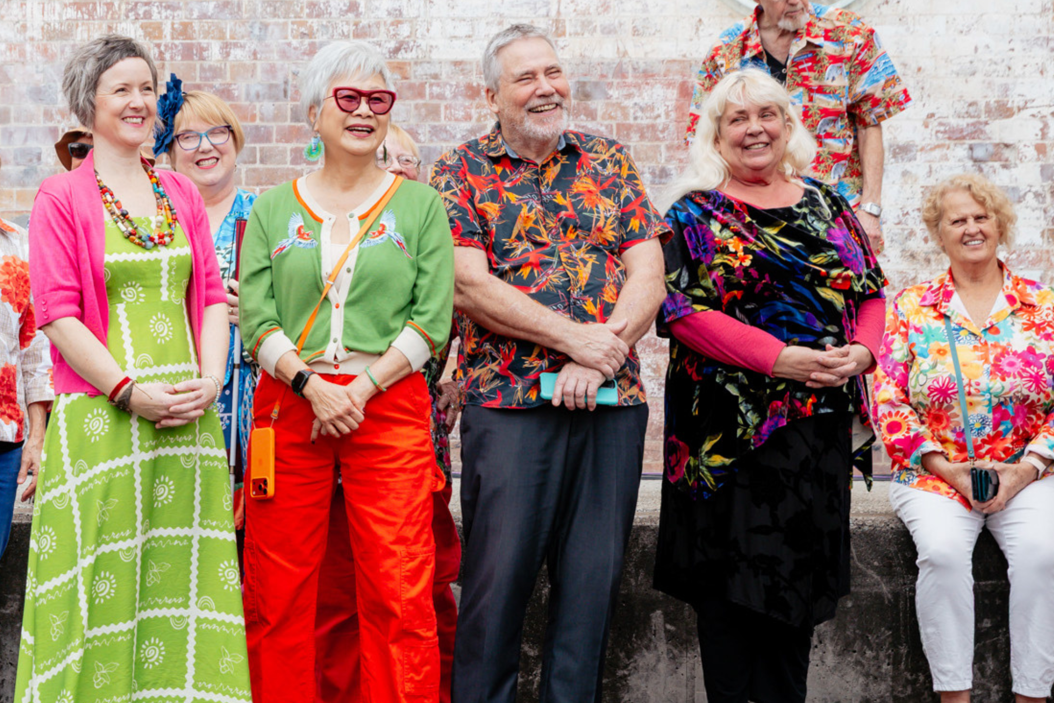 A group of older adults stand outdoors, smiling and laughing. They wear colorful, vibrant clothing, with bold patterns and bright accessories, against a textured brick wall background.