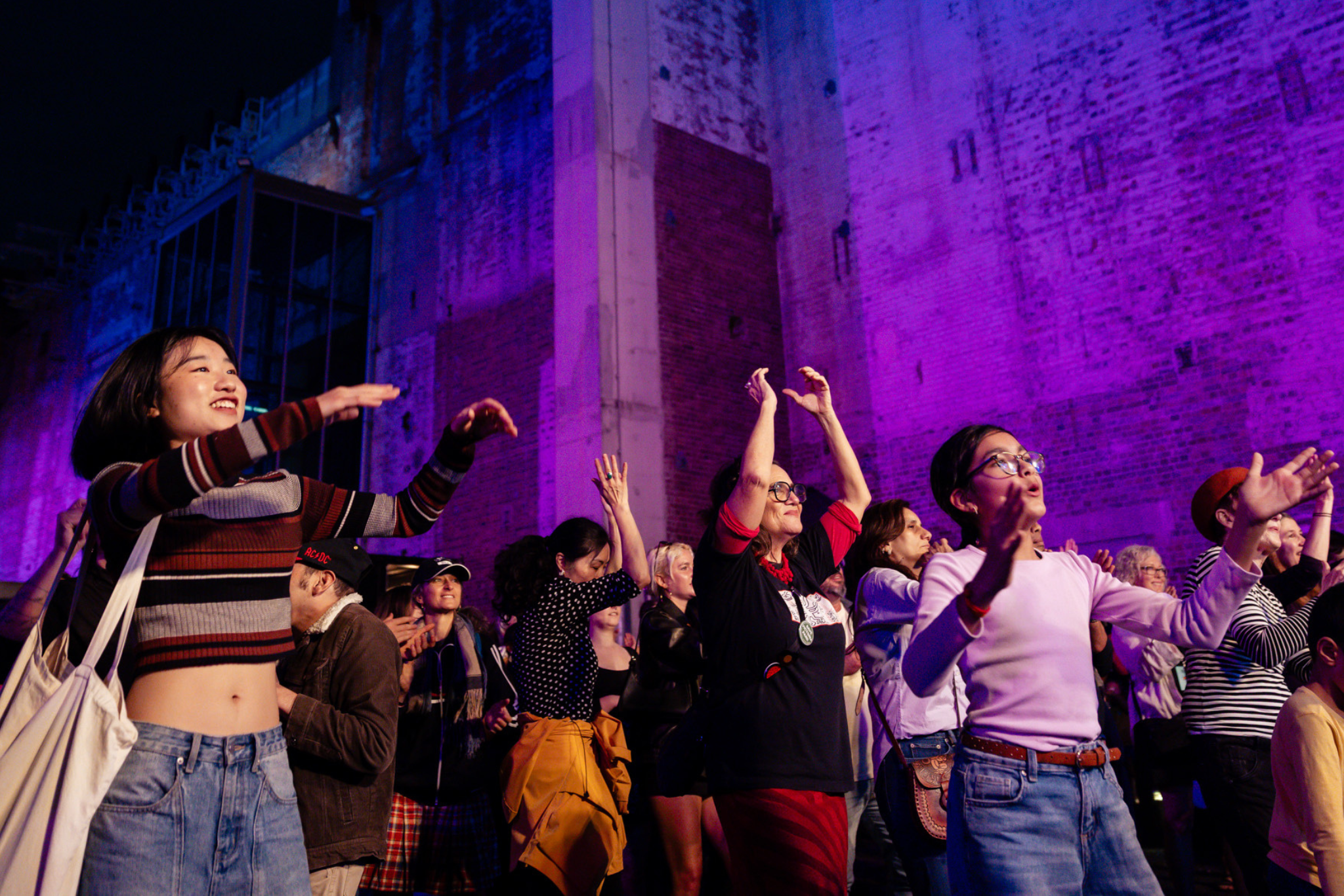 A crowd of people dances joyfully outdoors at night in front of a large, illuminated brick wall washed in purple and blue light. Many attendees have their arms raised and are smiling.