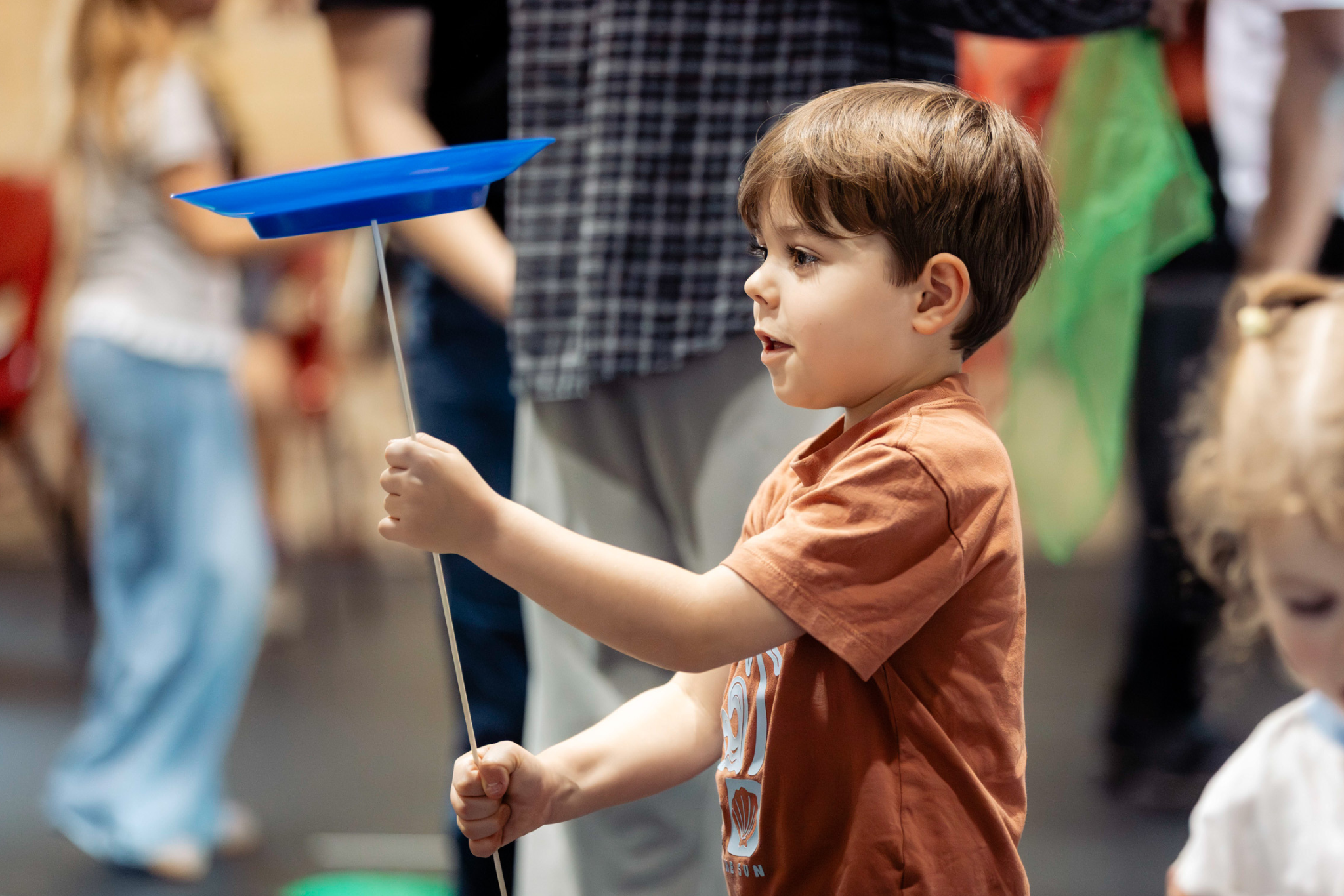 A young boy in a brown shirt spins a blue plate on a stick, focusing intently. Other people and colorful objects are blurred in the background, suggesting an indoor event or activity.
