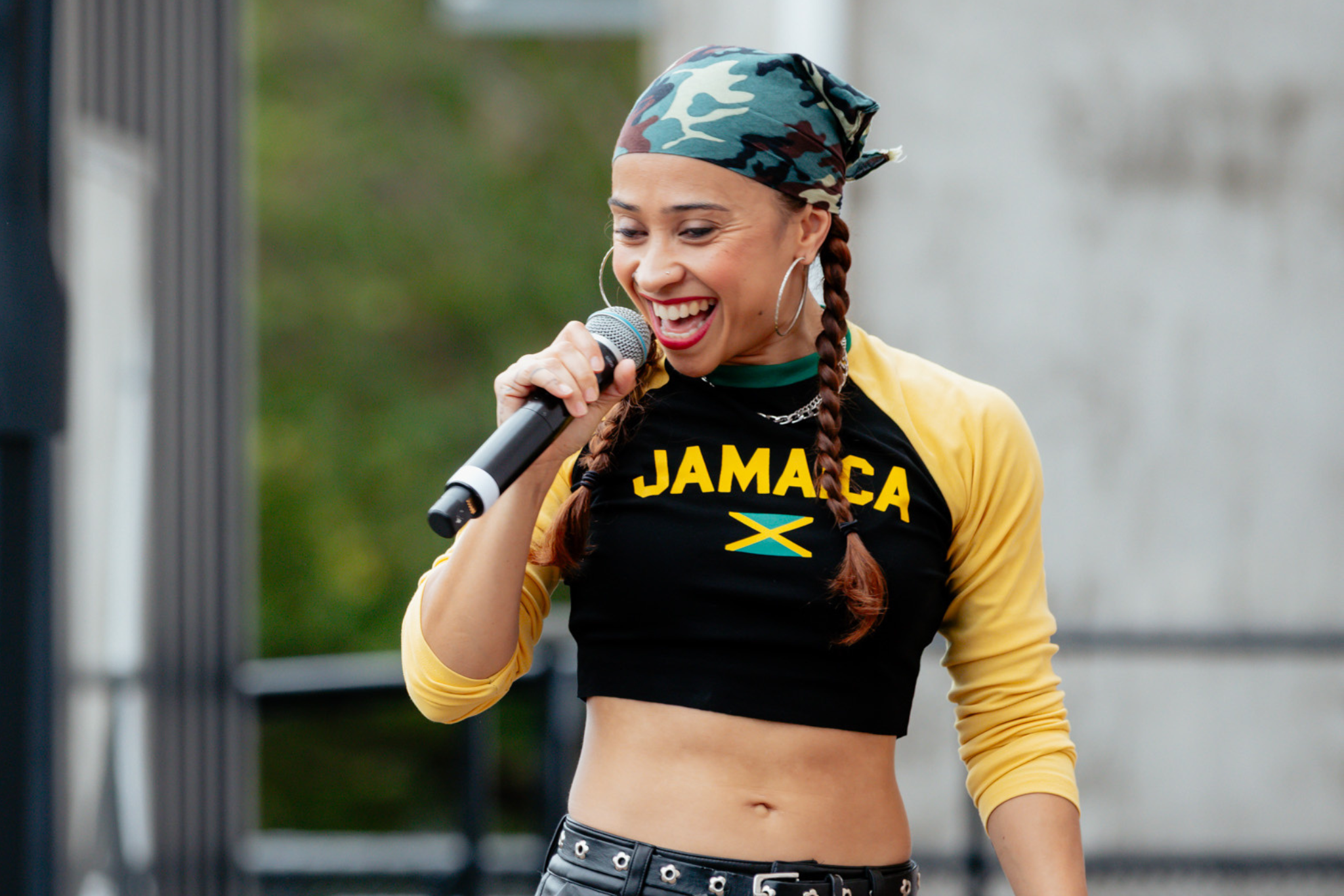 A smiling woman with braids, wearing a Jamaica crop top and a camo bandana, holds a microphone while performing outdoors.