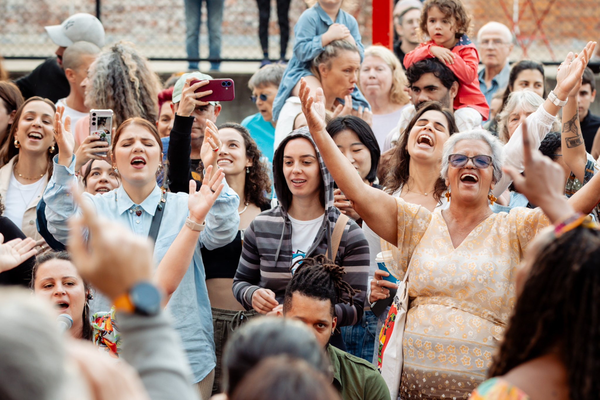 A diverse crowd of people outdoors, many smiling, raising their arms, and cheering. Some hold phones to record the moment, while others clap or sing along, creating an energetic, joyful atmosphere.
