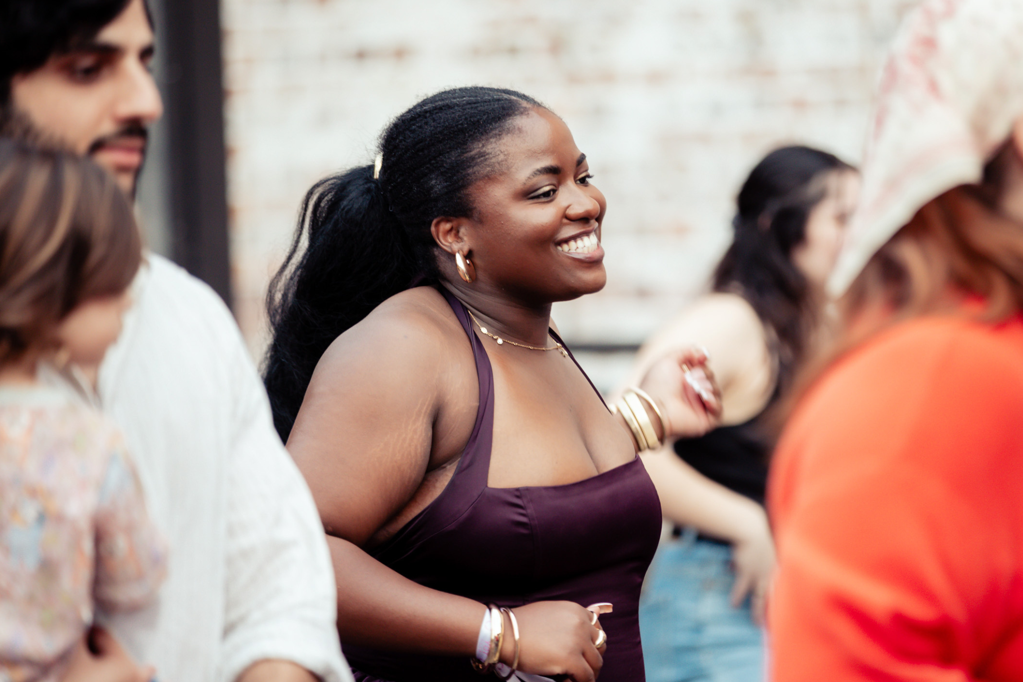 A woman wearing a sleeveless dress smiles brightly while dancing outdoors among a group of people.