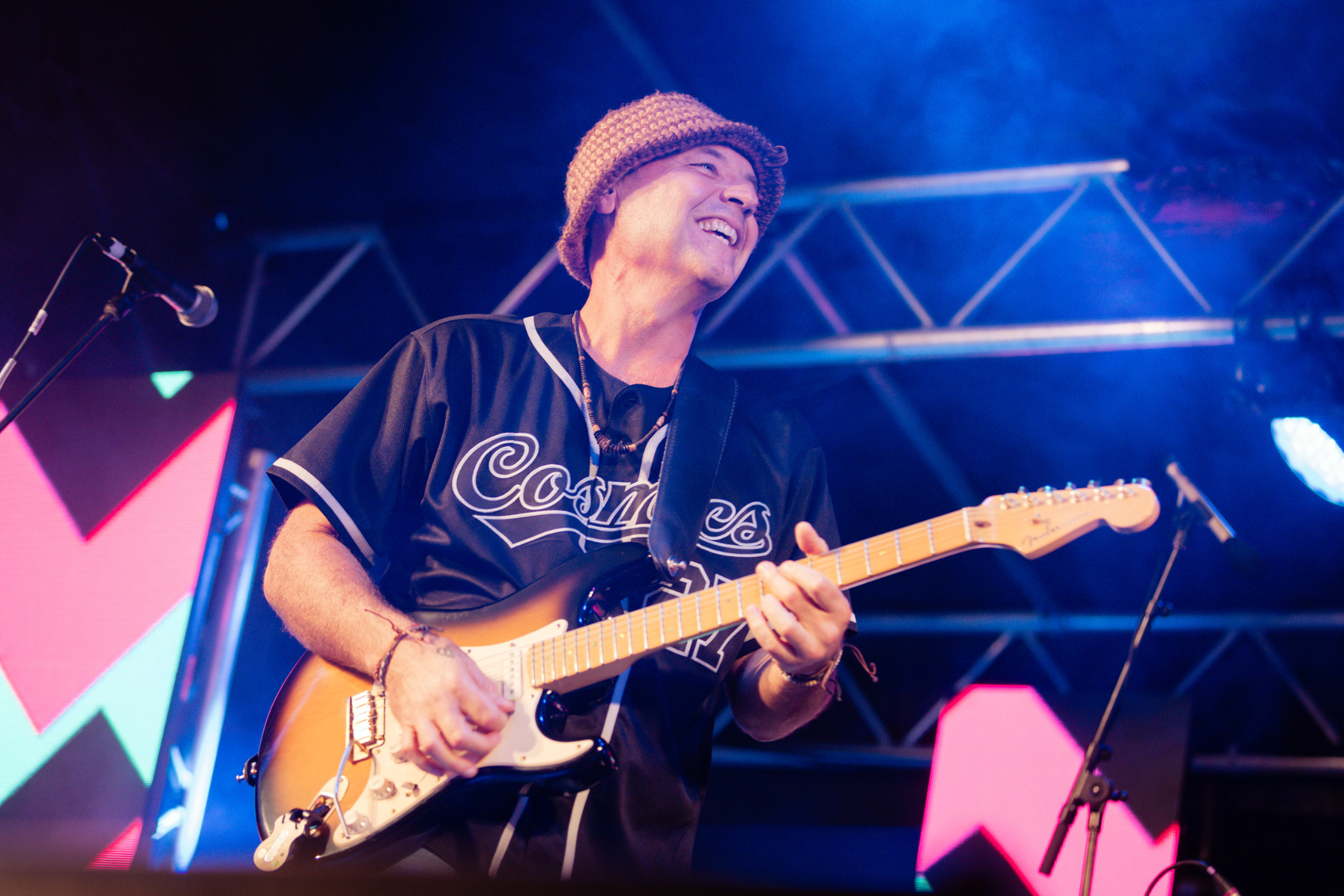 A musician wearing a knit hat and Cosmos jersey plays an electric guitar and smiles on stage, with colorful lights and abstract shapes in the background.