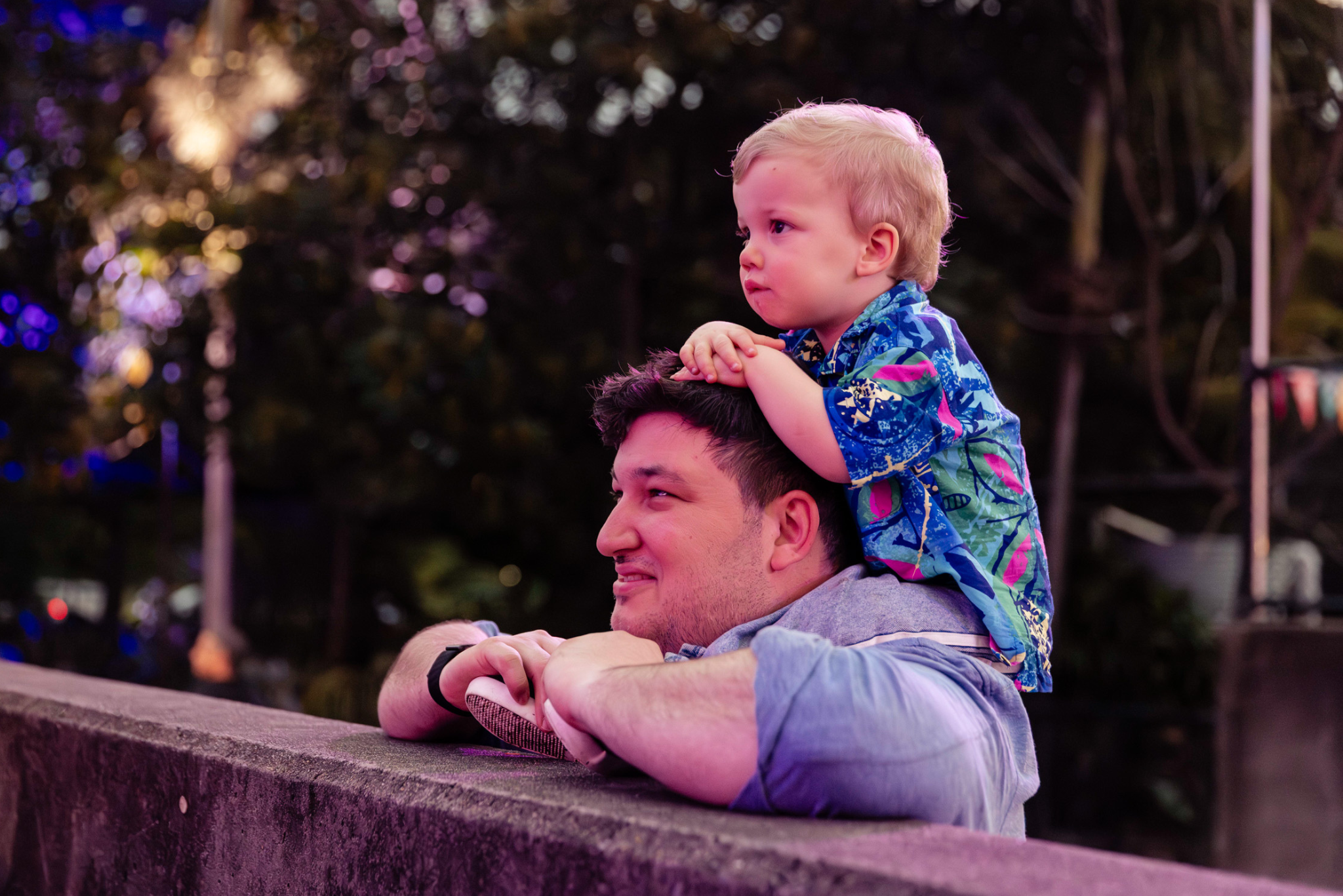 A man leans on a stone railing, smiling, while a young child in a colorful shirt sits on his shoulders, looking ahead. They are outdoors at dusk, surrounded by trees and soft purple lighting.