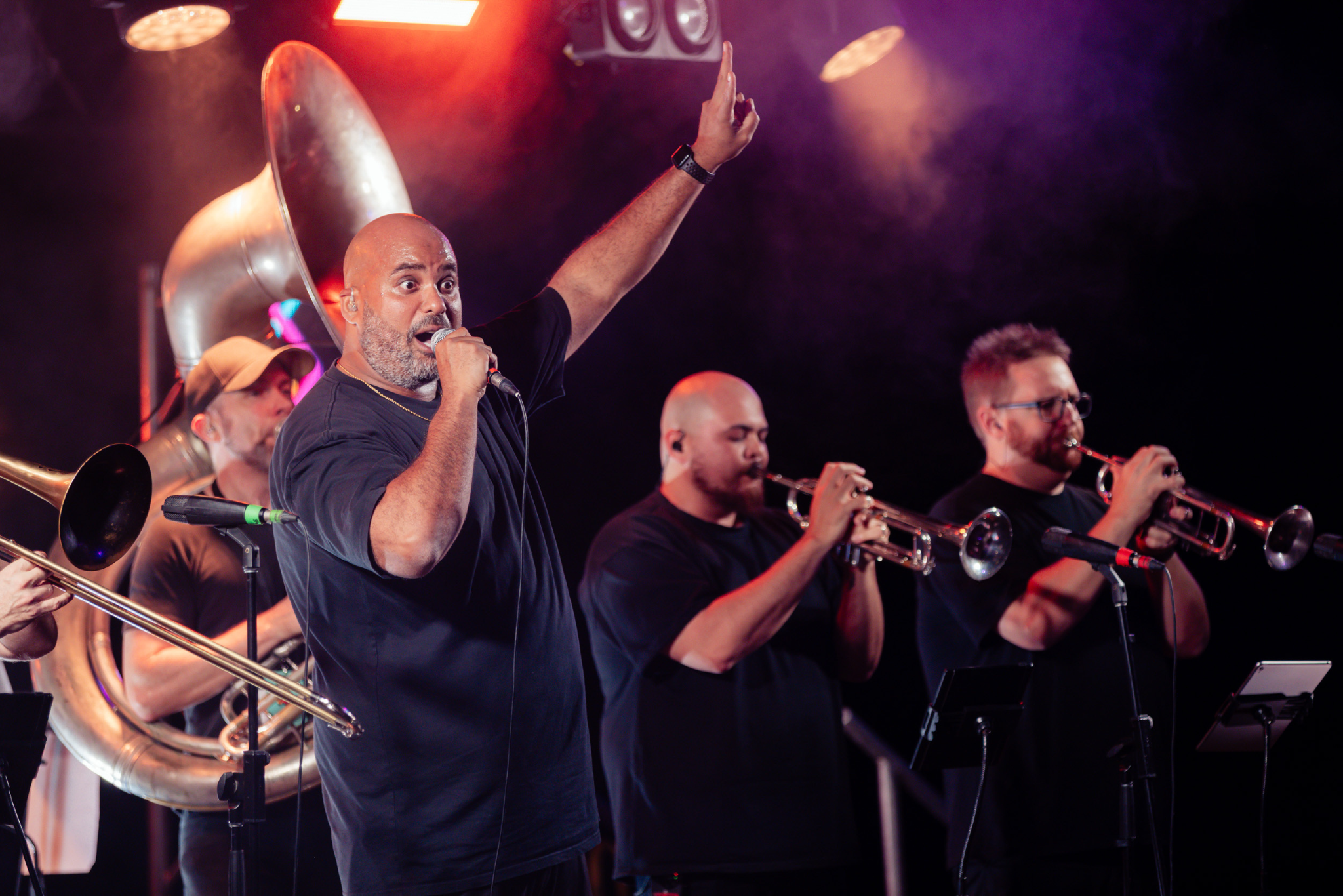 A man sings passionately into a microphone with one arm raised as musicians beside him play brass instruments on a stage under colorful lights.