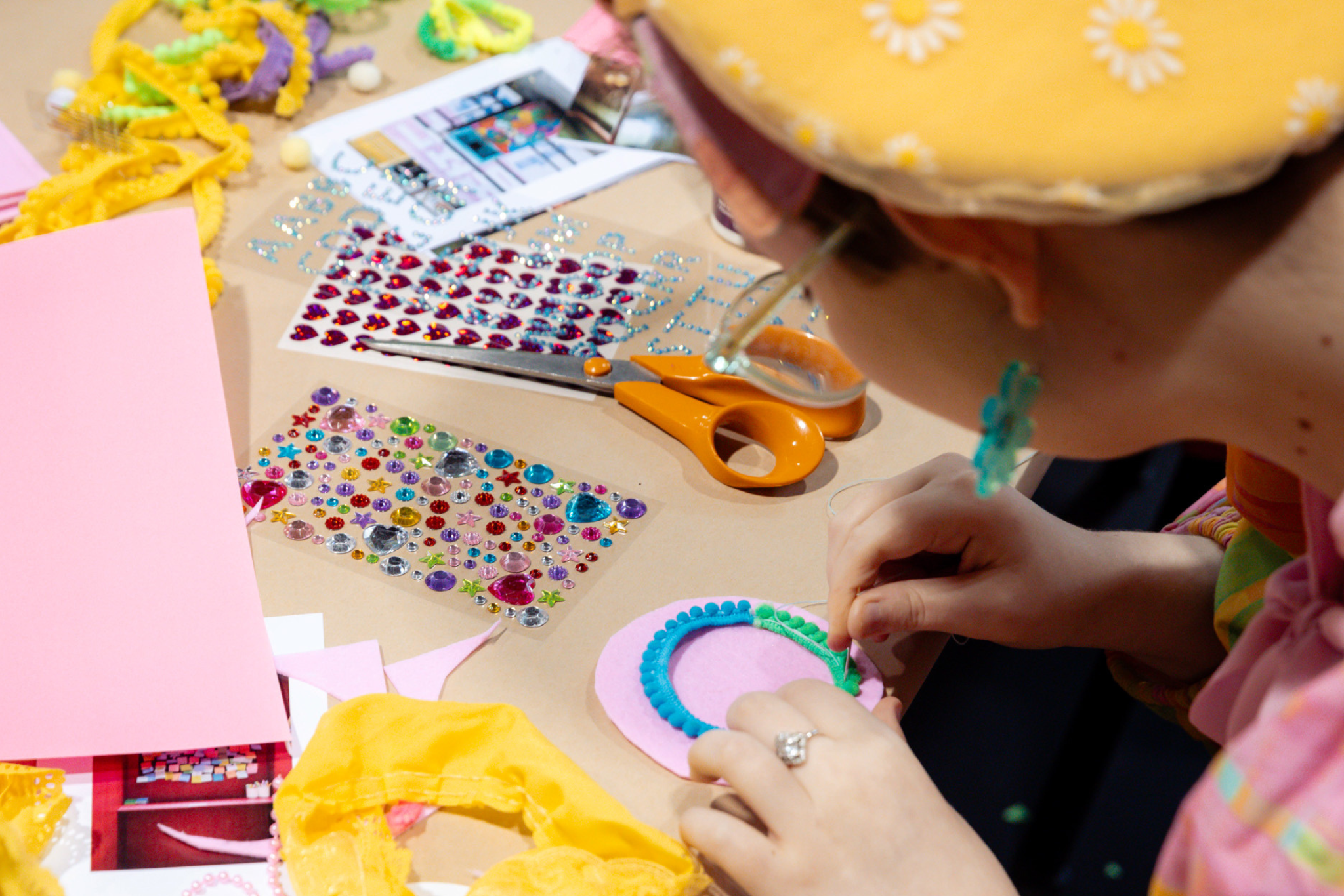 A person decorates a pink paper ring with colorful beads and gems on a table filled with craft supplies, scissors, and stickers.
