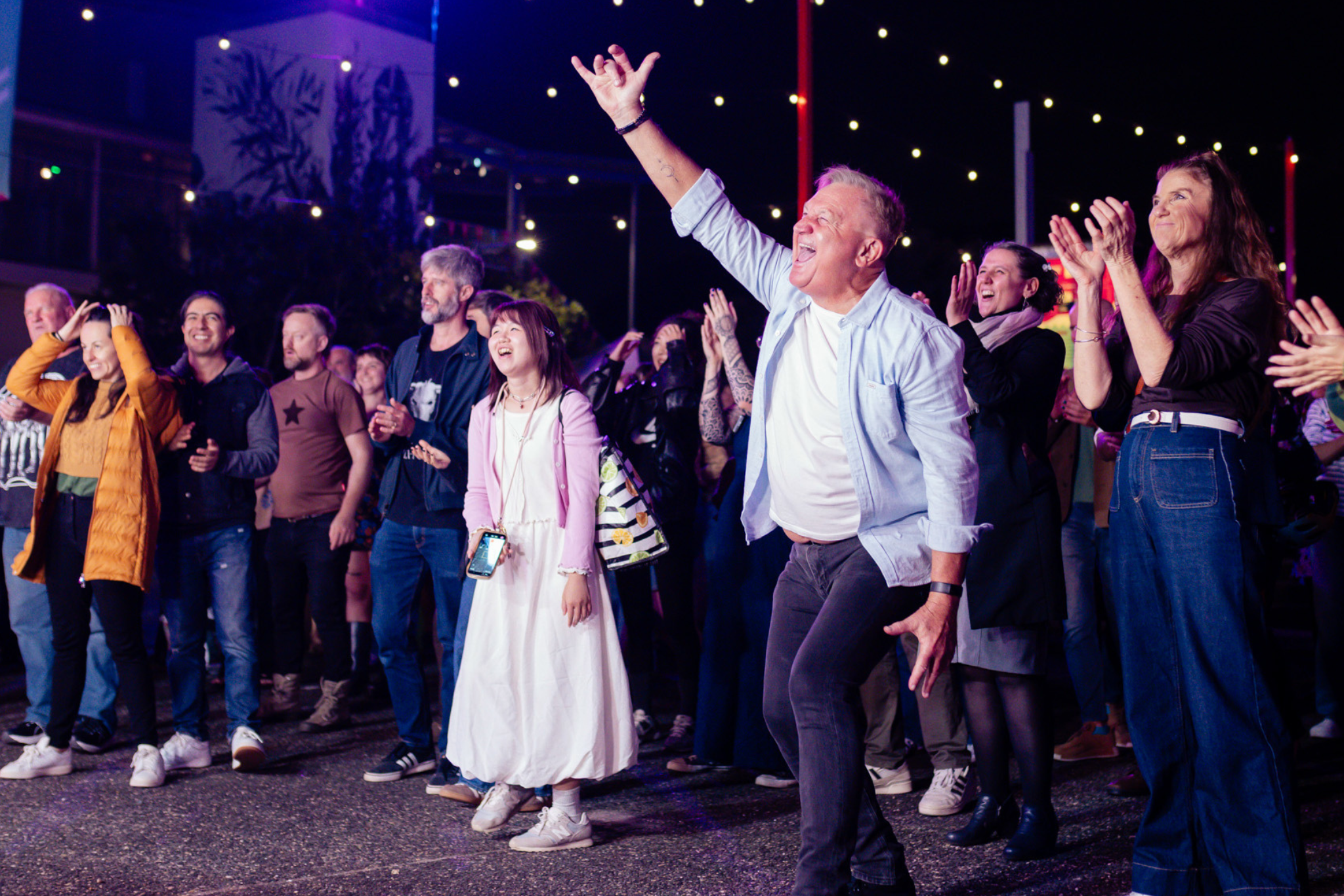 A group of people enjoying an outdoor concert at night; one man stands out mid-cheer with his arm raised, while others clap, smile, and watch the performance under string lights.