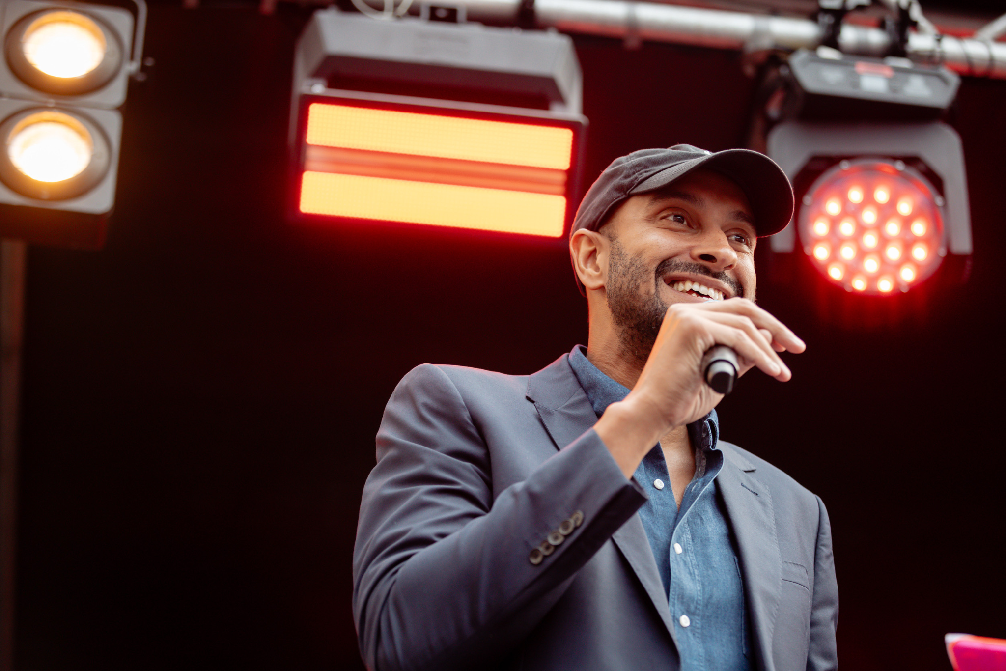 A man in a black cap and blazer smiles while speaking into a microphone on stage, with red stage lights shining behind him.