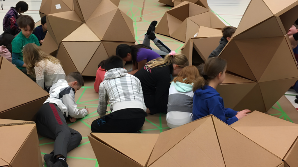 Children sit and kneel on the floor, drawing or writing on large geometric cardboard structures in a gymnasium-like setting.