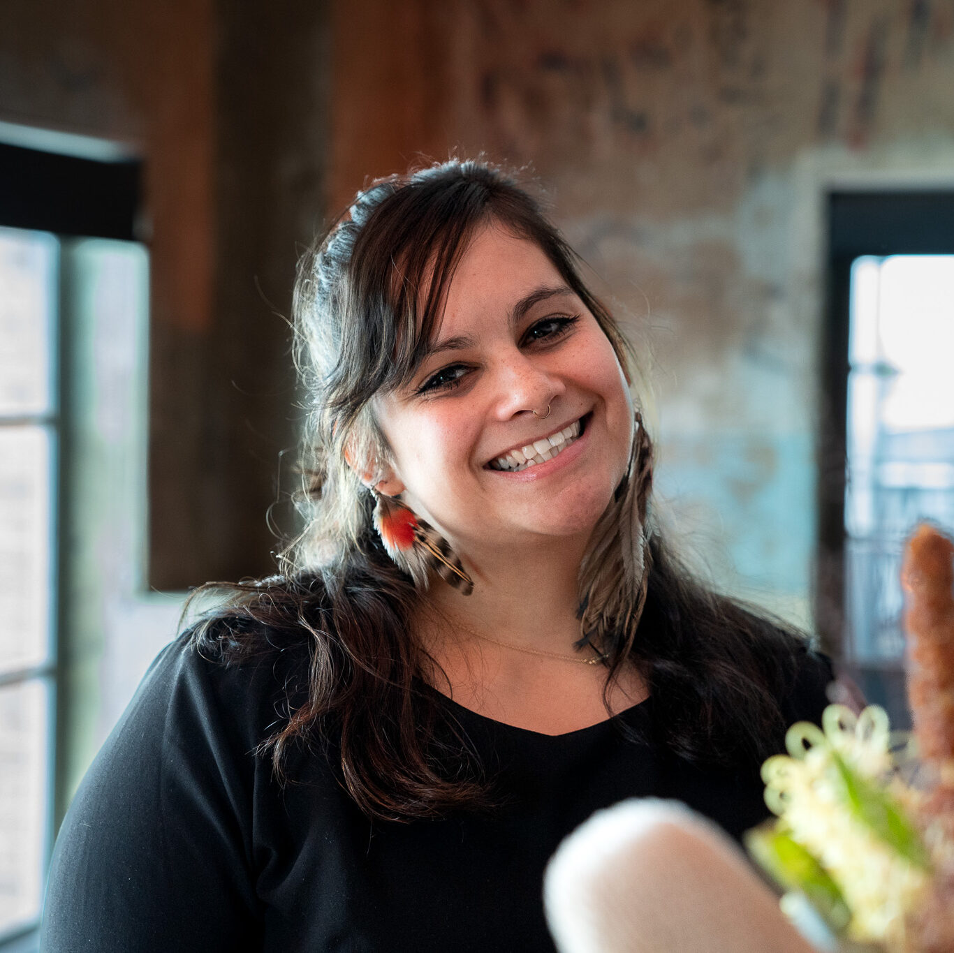 A woman with long dark hair wearing a black top smiles warmly indoors, with blurred windows and rustic decor in the background.