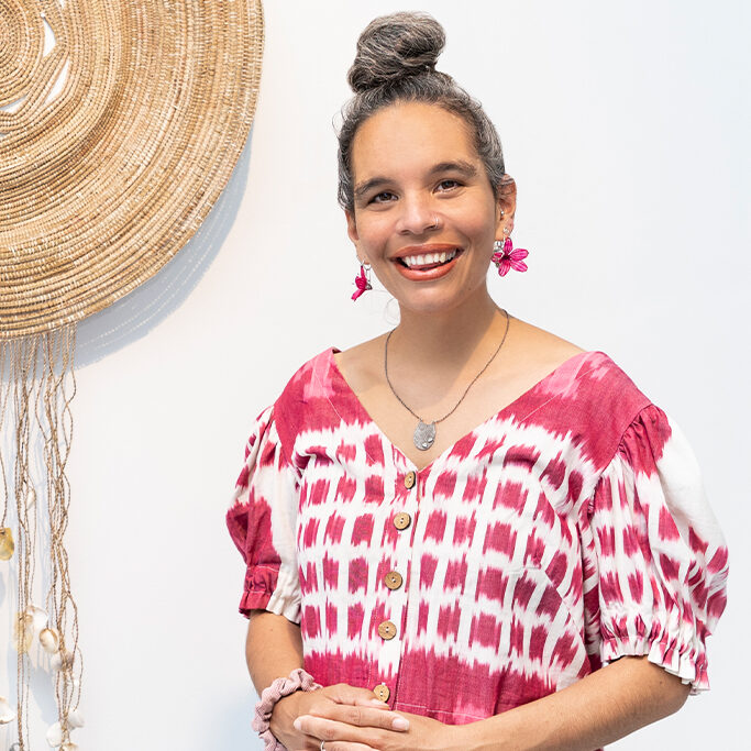 A woman with gray hair in a bun, wearing a red and white patterned blouse and blue jeans, stands smiling in front of a white wall with a large woven decoration hanging beside her.