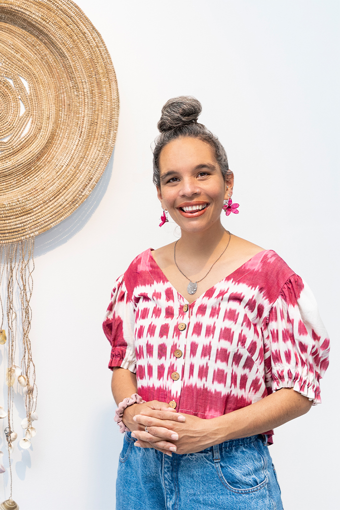 A woman with gray hair in a bun, wearing a red and white patterned blouse and blue jeans, stands smiling in front of a white wall with a large woven decoration hanging beside her.