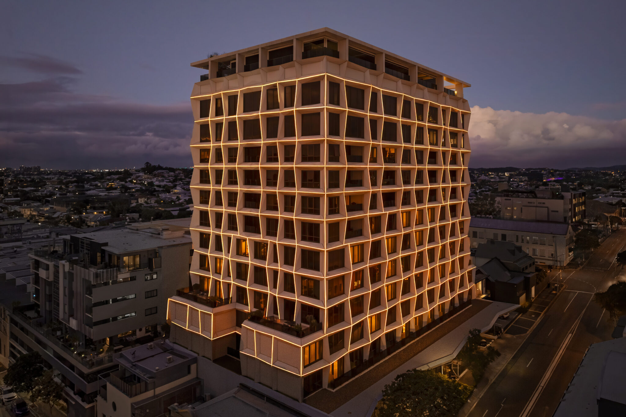 A modern multi-story building with a geometric, grid-like facade is illuminated with warm yellow lights in an urban area at dusk—inviting you to experience vibrant city life reminiscent of cultural hubs like Visit Brisbane Powerhouse.