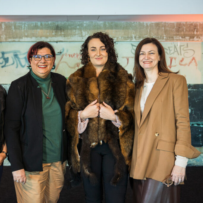 Three women stand together smiling. The woman in the center wears a brown fur wrap, while the others are in blazers. They stand indoors in front of a wall with faded graffiti.