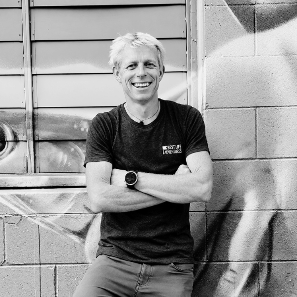 A smiling man with his arms crossed leans against a graffiti-covered wall, wearing a t-shirt that says Best Life Adventures.