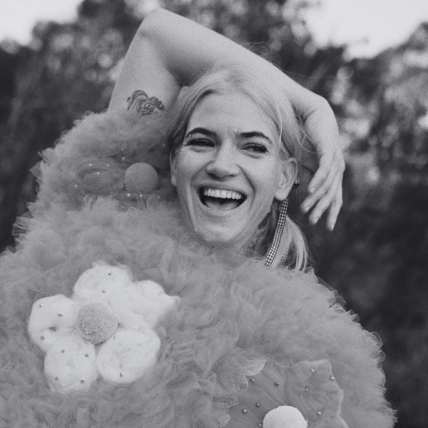 A person laughing with one arm raised behind their head, wearing a textured garment with large floral appliqués.
