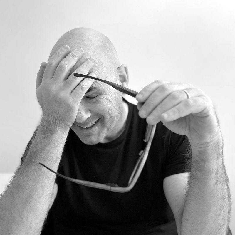 A smiling bald man with his hand over his forehead, holding a pair of glasses.